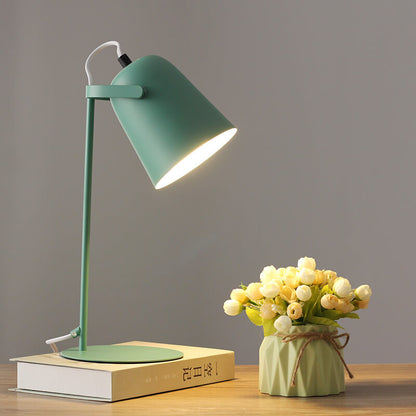 Grön bordslampa med tänd skärm på en bok, bredvid en ljusgrön kruka med blommor. 9