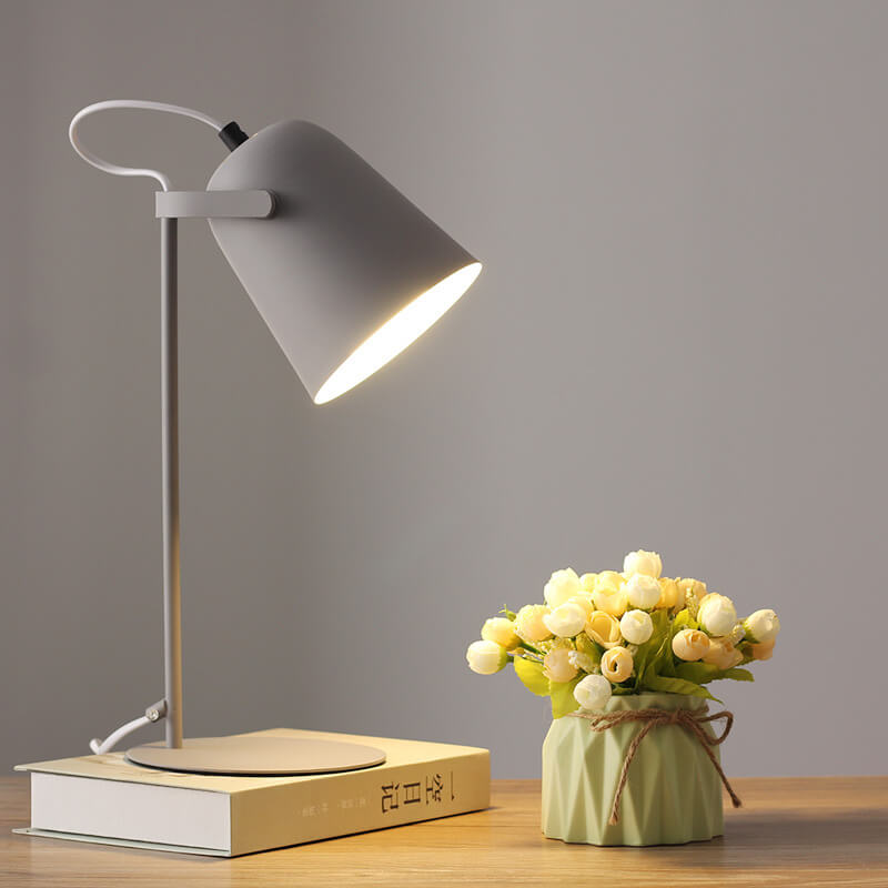 Grå bordslampa med tänd skärm på en bok, bredvid en ljusgrön kruka med vita och gula blommor. 10