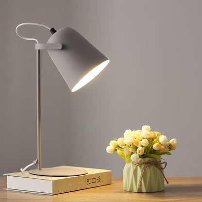 Grå bordslampa med tänd skärm på en bok, bredvid en ljusgrön kruka med vita och gula blommor. 10