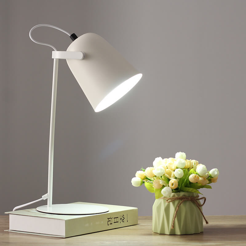 Vit bordslampa med tänd skärm på en bok, bredvid en grön kruka med vita och gula blommor. 11