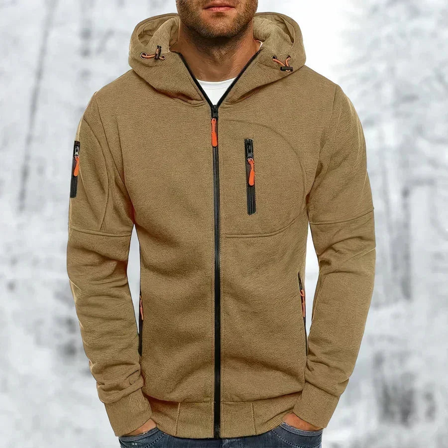 Man i beige-brun huvtröja med dragkedja och orange dragkedjehandtag, framifrån med händerna i fickorna, snöig bakgrund. 10
