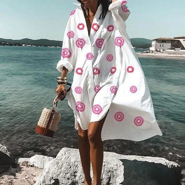 Frau im weißen Sommerkleid mit rosa Kreisen am Strand, hält eine geflochtene Handtasche.