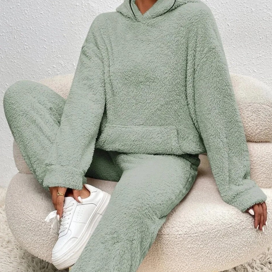 Frau in grünem Fleece-Hoodie und Jogginghose auf Sofa, weiße Sneaker, gemütliche Mode.