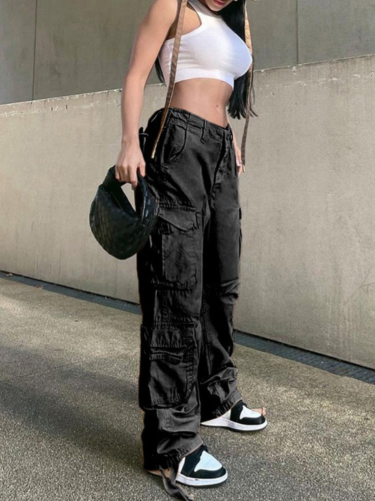Frau in schwarzer Cargohose, weißem Crop-Top, Sneakern, hält schwarze Handtasche.