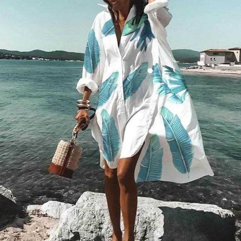 Frau im weißen Strandkleid mit blauen Blättern, hält geflochtene Tasche, am Meer.
