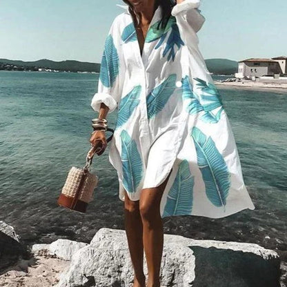 Frau im weißen Strandkleid mit blauen Blättern, hält geflochtene Tasche, am Meer.