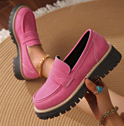 Rosa loafers med tjock svart sula, den ena skon hållen i handen medan modellen sitter på en flätad matta. 18