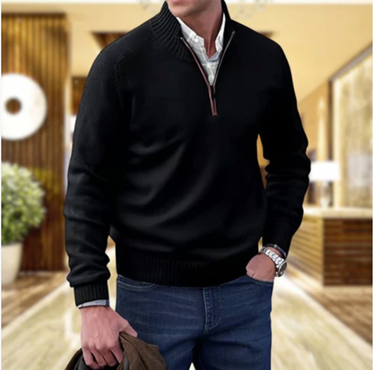 Mann in schwarzem Pullover mit Reißverschluss, blaue Jeans, moderne Inneneinrichtung im Hintergrund.