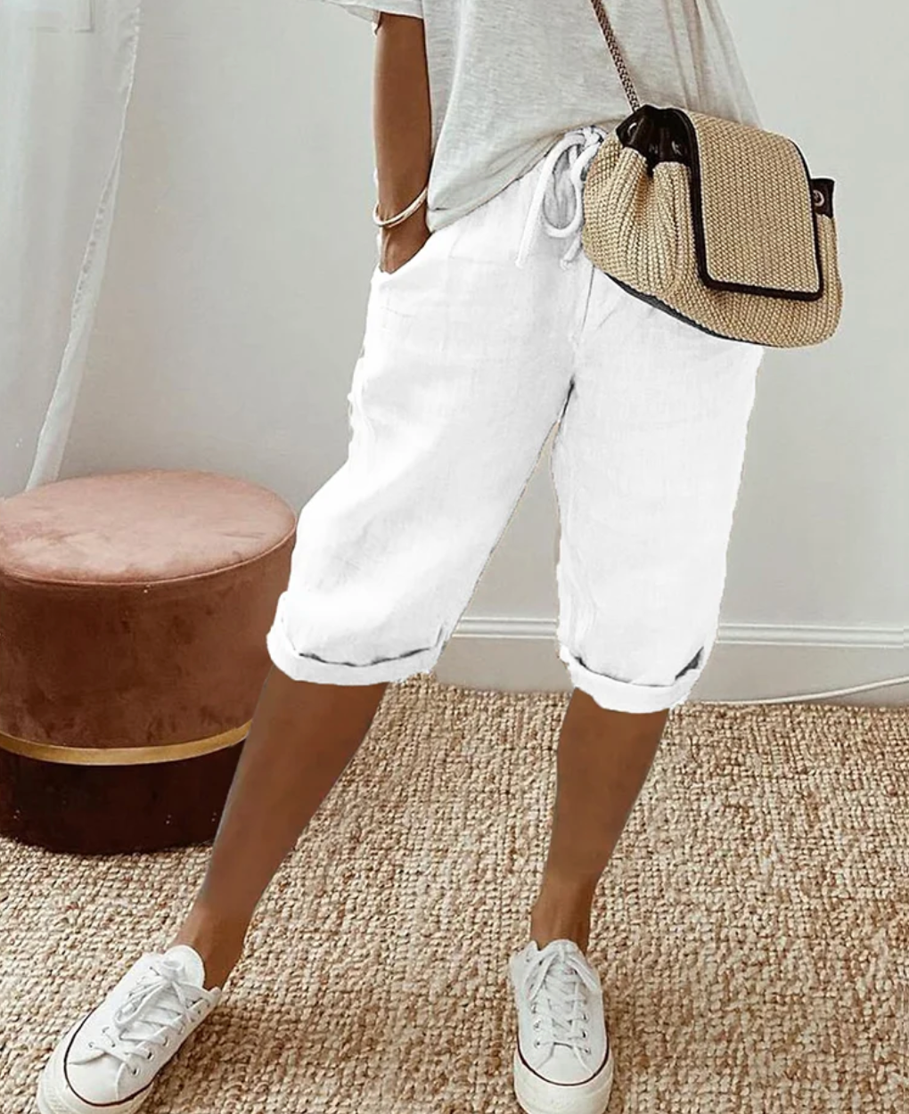 Frau in weißer Leinenhose, grauem T-Shirt, Strohtasche, weiße Sneaker, lässiger Sommerlook.
