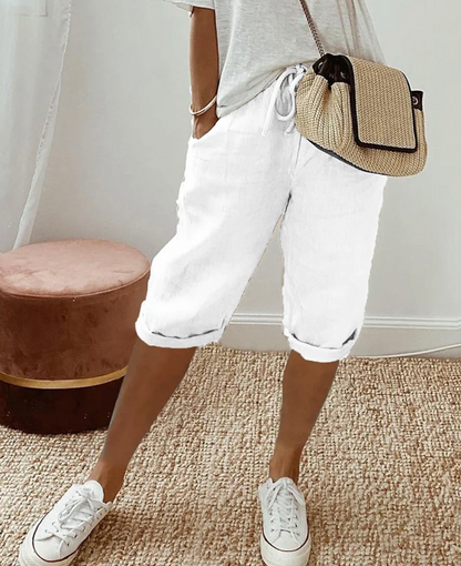 Frau in weißer Leinenhose, grauem T-Shirt, Strohtasche, weiße Sneaker, lässiger Sommerlook.