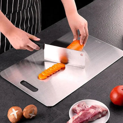 Person skivar en morot med en bred kökskniv på metallskärbrädan, med kött, tomat och svamp i bakgrunden. 3