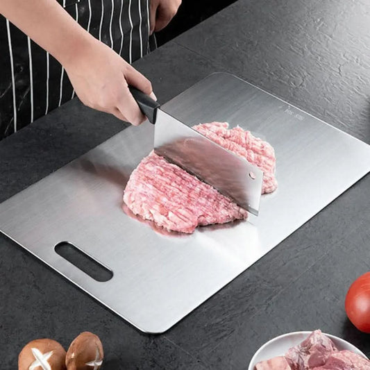 Metallskärbräda på en mörk bänk där en person hackar rött köttfärs med en kökskniv, bredvid svamp och tomat. 1