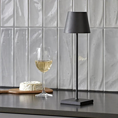 Schwarze Tischlampe aus Metall neben Weinglas und Käse auf grauem Küchentresen vor Fliesenwand.