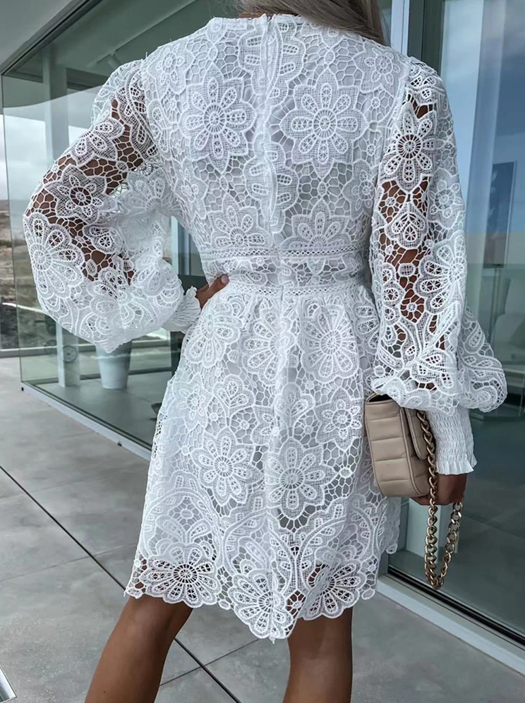 Frau in weißem Spitzenkleid mit langen Ärmeln, beige Tasche, Balkonhintergrund.