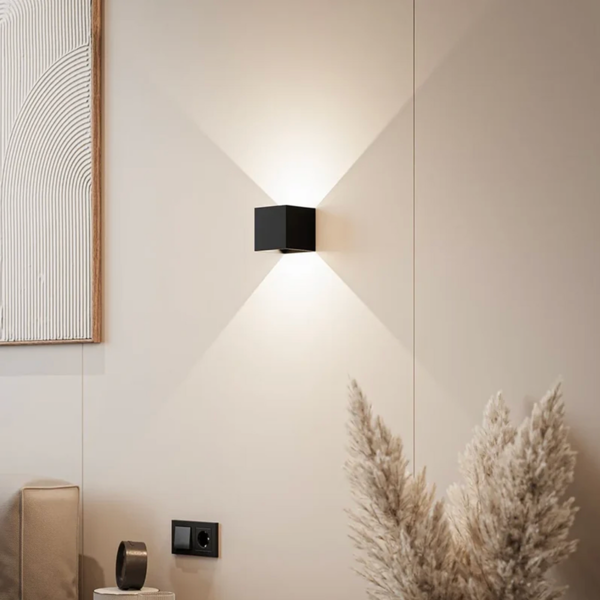 Svart kubformad vägglampa som lyser uppåt och nedåt på en beige vägg i ett vardagsrum med pampasgräs och tavla. 1