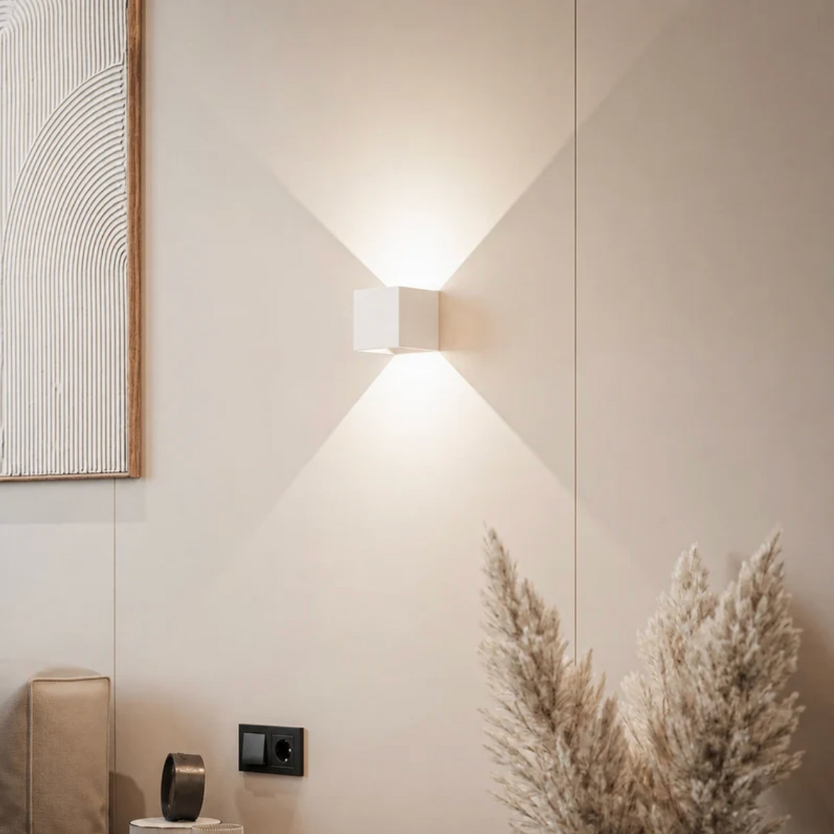 Vit kubformad vägglampa på en beige vägg som sprider ljus upp och ner, med pampasgräs och tavla i bilden. 12