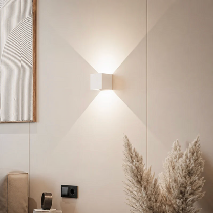 Vit kubformad vägglampa på en beige vägg som sprider ljus upp och ner, med pampasgräs och tavla i bilden. 12