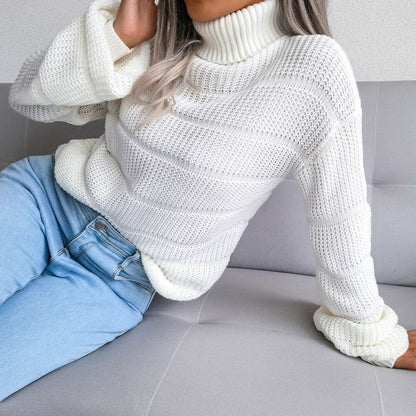 Frau in weißem Strickpullover mit Rollkragen und blauer Jeans auf grauem Sofa.