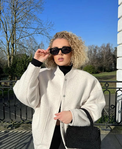 Frau mit weißer Wolljacke, schwarzer Sonnenbrille und Tasche im Freien bei sonnigem Wetter.