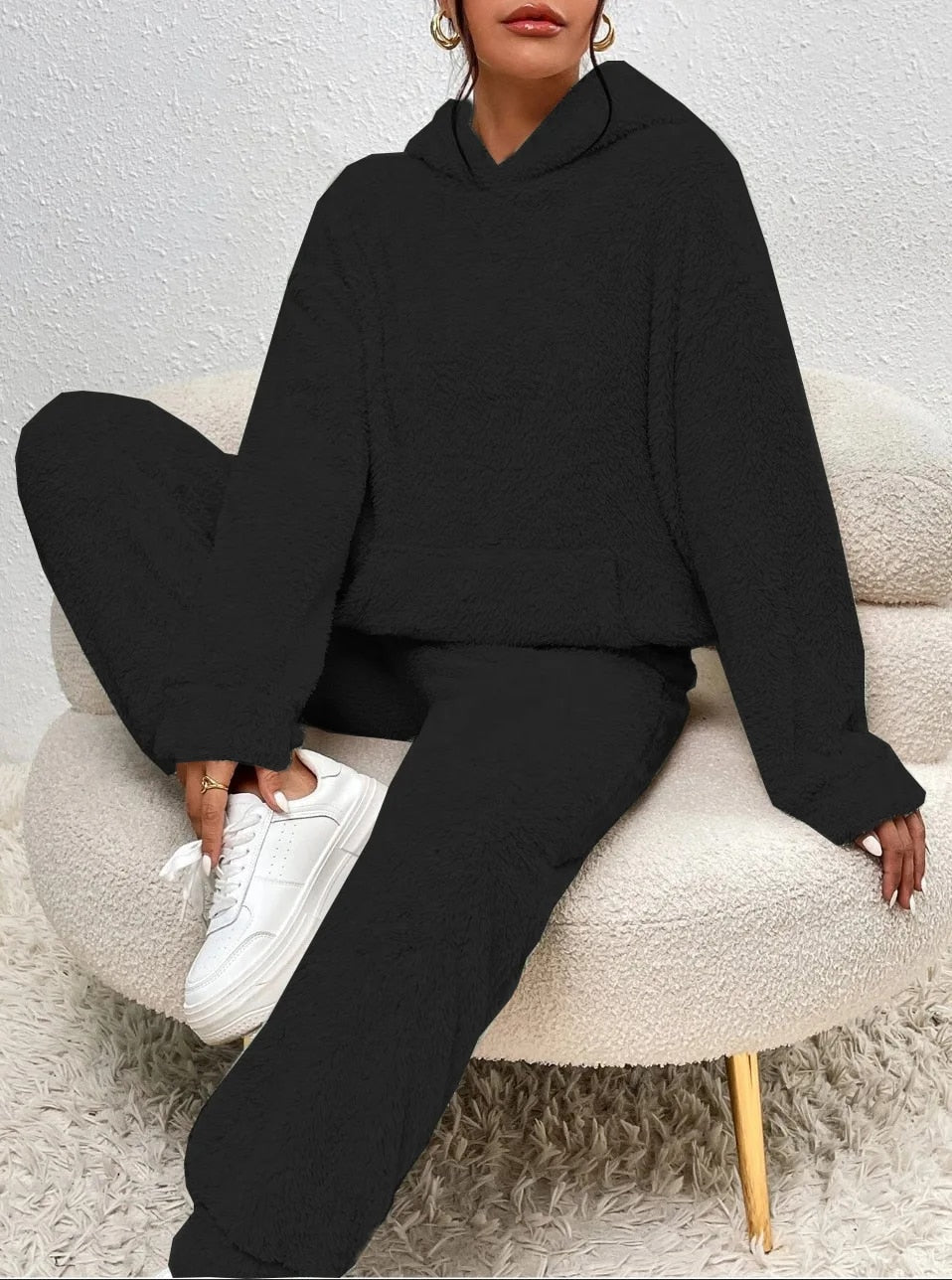 Frau in schwarzem Fleece-Hoodie und Jogginghose, weiße Sneaker, sitzt auf modernem Sessel.
