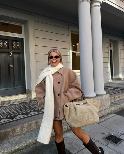 Frau in beiger Jacke mit weißem Schal und großer Tasche vor Gebäude mit Säulen.