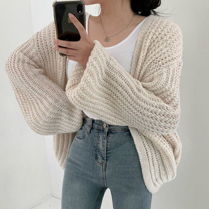 Frau in cremefarbenem Strickcardigan, weißem T-Shirt und blauer Jeans, Smartphone in der Hand.