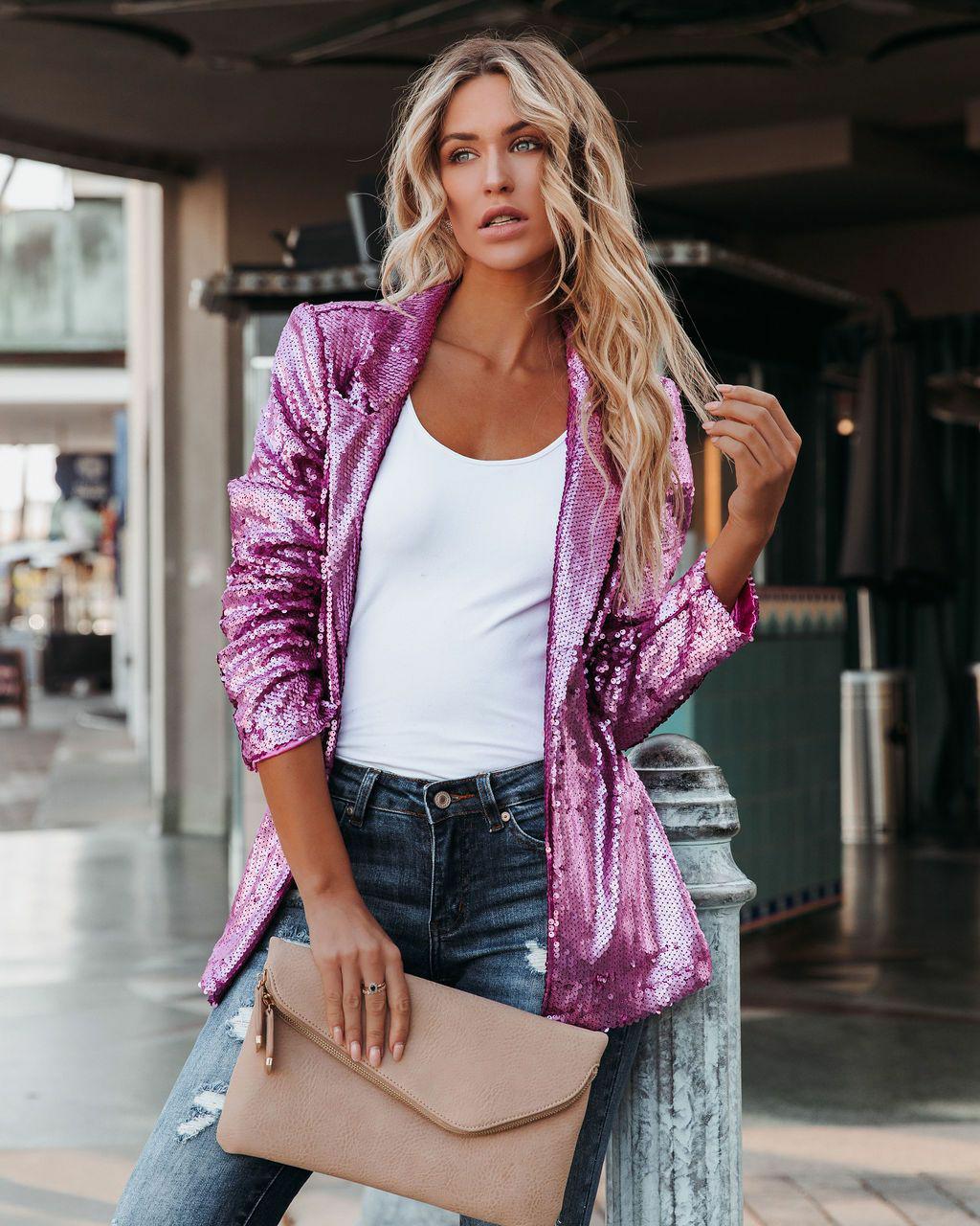 Frau in pinkem Paillettenblazer, weißem Top, Jeans, hält beige Clutch, modisch, urban.
