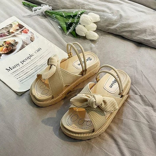 Beige Sandalen mit Schleife, Damen, Sommermode, bequem, flach, auf grauem Bett, Tulpen im Hintergrund.