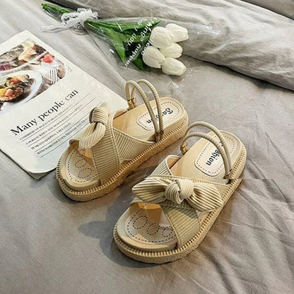 Beige Sandalen mit Schleife, Damen, Sommermode, bequem, flach, auf grauem Bett, Tulpen im Hintergrund.