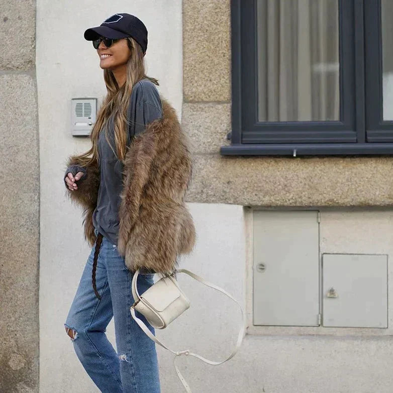 Frau in brauner Kunstpelzjacke, blauer Jeans und schwarzer Kappe mit weißer Umhängetasche.