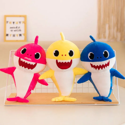 Drei bunte Baby Shark Plüschtiere, rosa, gelb, blau, auf einem Tisch stehend.