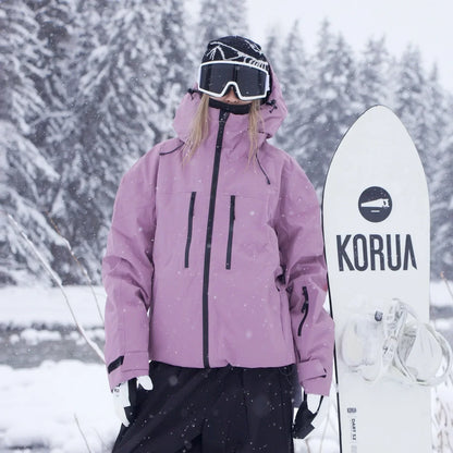 Modell i rosa jacka med svarta dragkedjor och justerbar huva står bredvid ett vitt snowboard i snöfall. 3