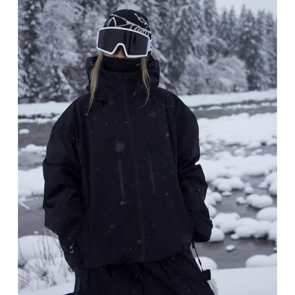 Modell i helsvart oversize-jacka med kragen uppfälld står vid en snötäckt bäck i snöfall. 2