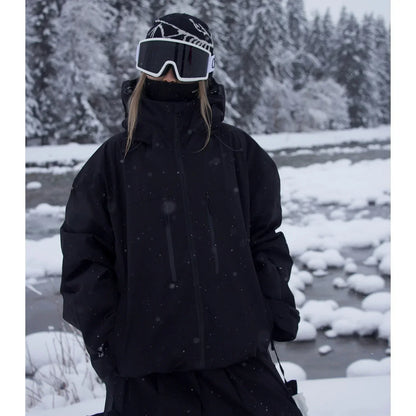 Modell i helsvart oversize-jacka med kragen uppfälld står vid en snötäckt bäck i snöfall. 2