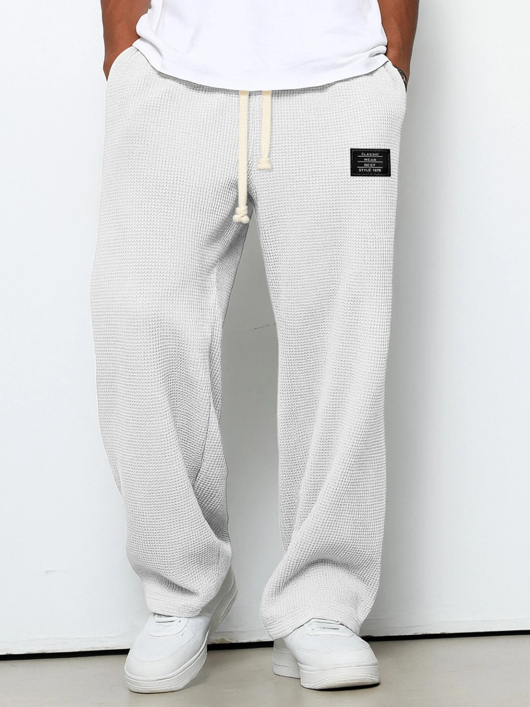 Man bär ljusgrå, nästan vita våffelstickade byxor med vida ben och dragsko, sedda framifrån med vit t-shirt och sneakers. 2
