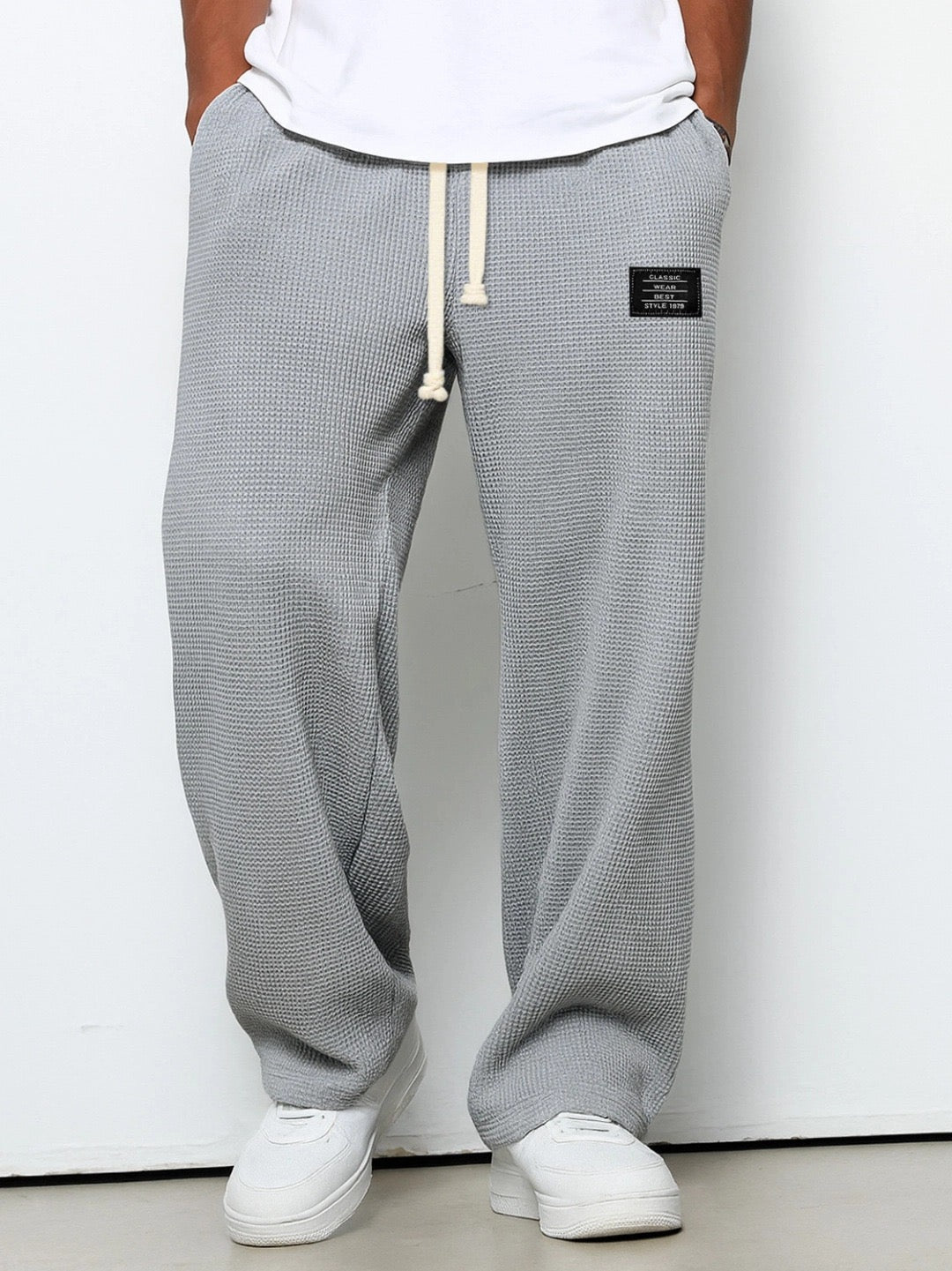 Man bär mellangrå våffelstickade byxor med vida ben och krämvit dragsko, framifrån med händerna i fickorna och vita sneakers. 3