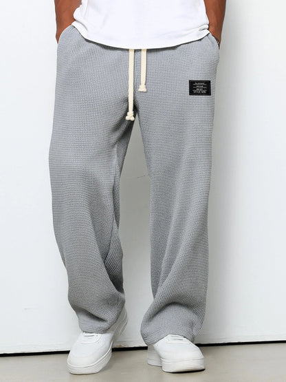 Man bär mellangrå våffelstickade byxor med vida ben och krämvit dragsko, framifrån med händerna i fickorna och vita sneakers. 3