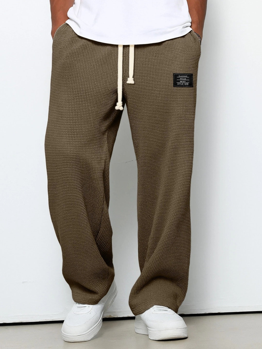Man bär bruna våffelstickade byxor med vida ben och krämvit dragsko, framifrån med händerna i fickorna och vita sneakers. 5