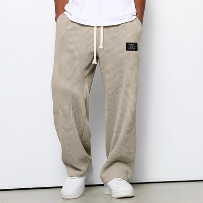 Man bär beige våffelstickade byxor med vida ben och krämvit dragsko, i närmare beskärning framifrån med vita sneakers. 6