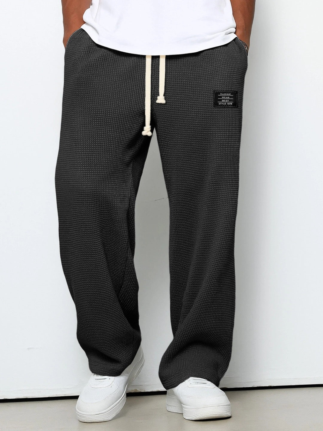 Man bär mörkgrå våffelstickade byxor med vida ben och krämvit dragsko, framifrån med händerna i fickorna och vita sneakers. 1
