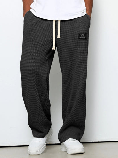 Man bär mörkgrå våffelstickade byxor med vida ben och krämvit dragsko, framifrån med händerna i fickorna och vita sneakers. 1