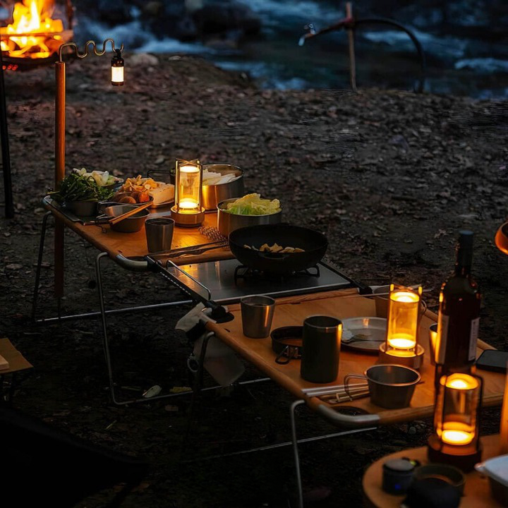 Tända lampor står på campingbord med kastruller och mat vid en eld på kvällen utomhus. 4