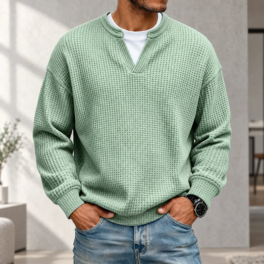 Man i mintgrön våffelstickad tröja med vit t-shirt under, händerna i jeansfickorna, ljus inomhusmiljö 1