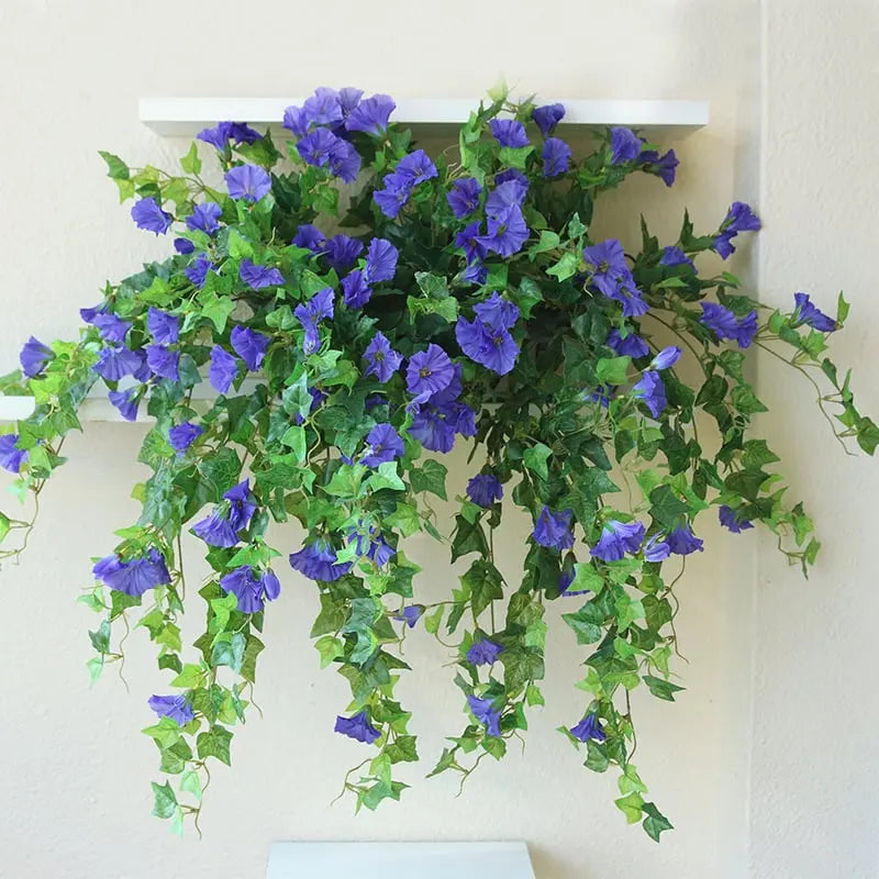 Lila blommor med gröna slingor hänger ned från en vit vägghylla mot en ljus vägg. 4