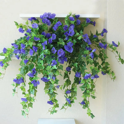 Lila blommor med gröna slingor hänger ned från en vit vägghylla mot en ljus vägg. 4