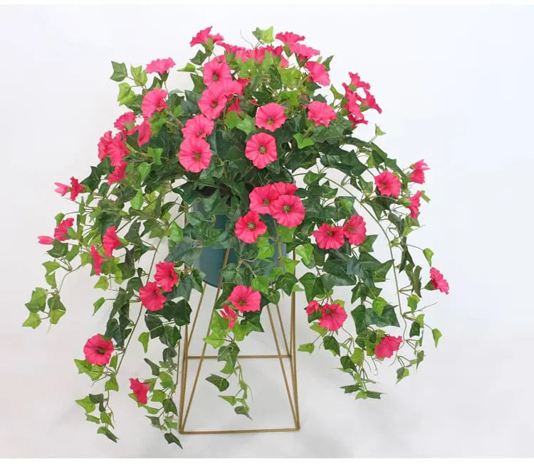 Rosa blommor och gröna slingrande blad hänger över kanten på en blågrön kruka på ett guldfärgat metallställ. 3