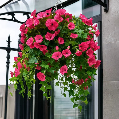 Rosa konstgjorda blommor med gröna blad i en hängande korg utomhus, vid en svart smidesgrind och en dörr. 1