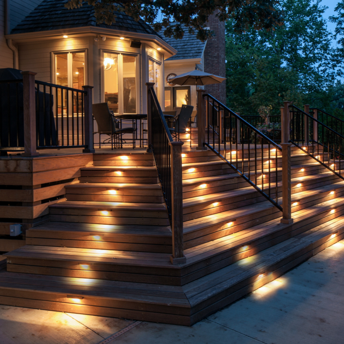 Beleuchtete Holzterrasse mit Treppen, LED-Lichtern, schwarzem Geländer, abends im Garten.