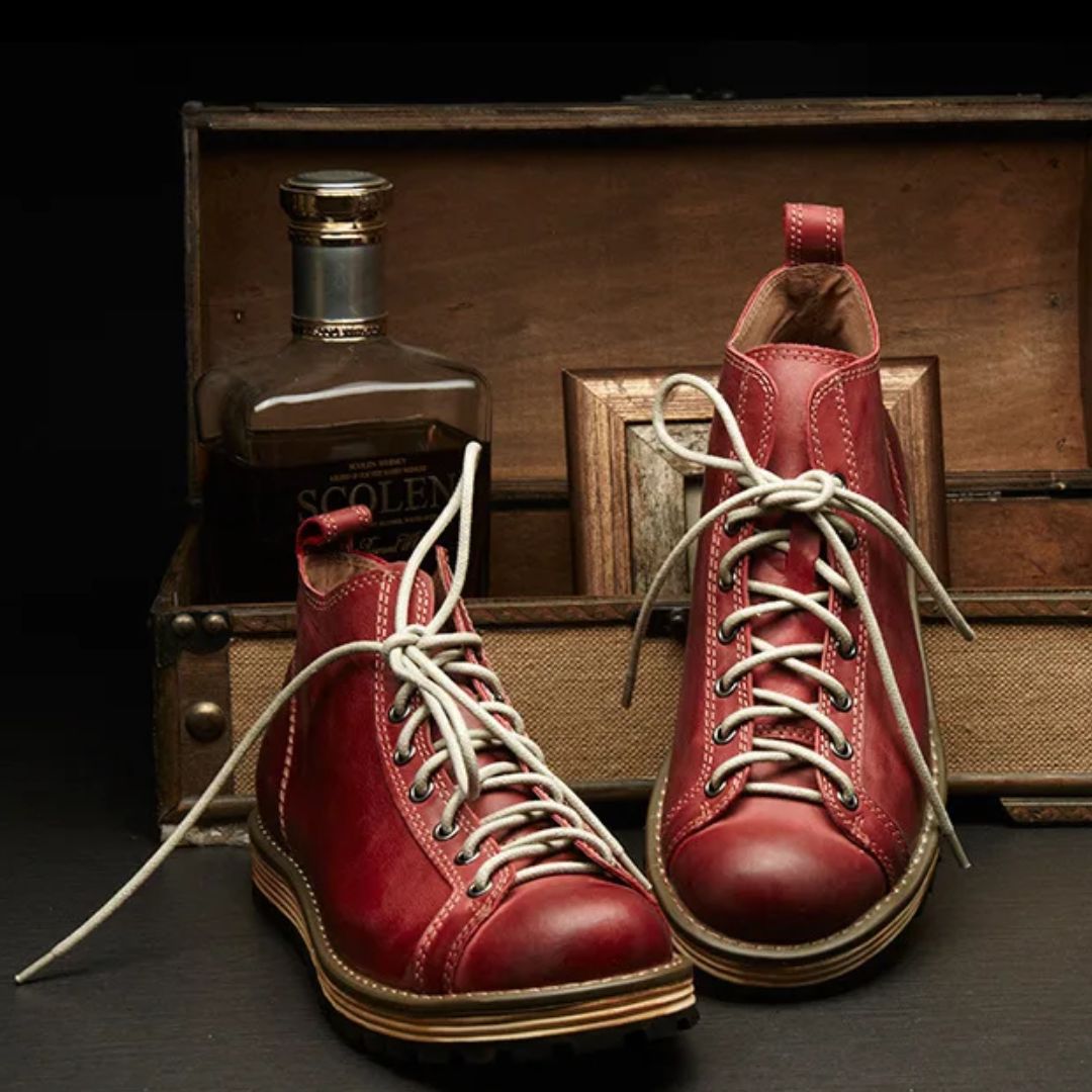 Rote Lederstiefel mit Schnürsenkeln vor Whiskyflasche und Holzkiste auf dunklem Hintergrund.