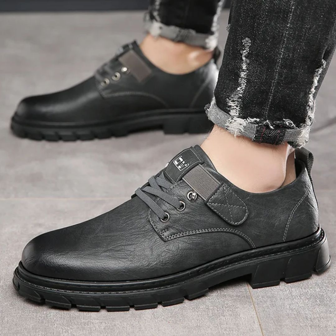 Schwarze Herren-Ledersneaker, lässig, robust, mit Schnürsenkeln, auf grauem Boden.